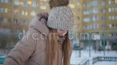 年轻女孩戴着冬帽，穿着柚子和<strong>羽绒服</strong>，在冬城的住宅建筑<strong>背景</strong>下行走。 快乐